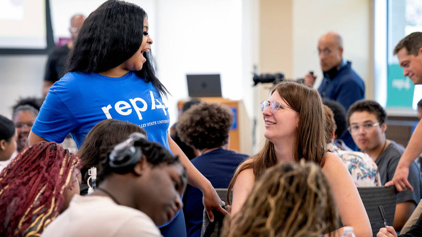 A student in a blue "rep4" shirt laughs while interacting with others in a classroom setting.